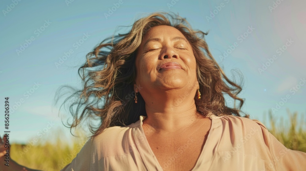 Wall mural Woman with closed eyes smiling in a field with wind-blown hair wearing a light-colored blouse and earrings with a blue sky and green grass in the background.