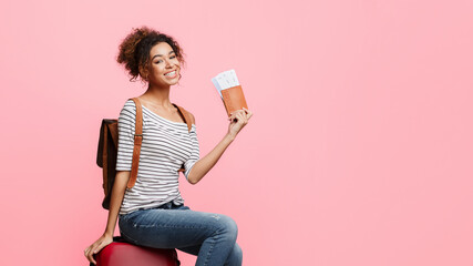 Excited african-american woman holding passport with tickets, sitting on suitcase, pink background