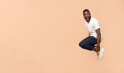 A young Black man with a white t-shirt and blue jeans jumps in the air against a peach-colored background. He is looking at the camera with a big smile and appears to be excited or happy.