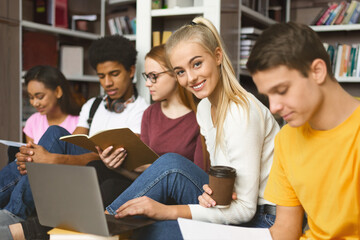 Focused international students sitting on wooden floor at library, getting ready for exams with books and laptops