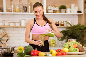 A woman in a pink sports bra pours a green smoothie from a pitcher into a glass in a kitchen.