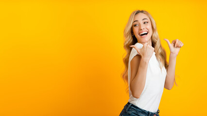 A young woman with long blonde hair smiles as she points with both thumbs towards the left side of the frame against a bright yellow background, panorama with copy space