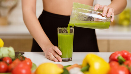 A woman in a black skirt pours a green smoothie into a glass from a pitcher, with fresh produce visible on the counter.