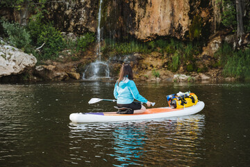 A woman sporting a blue hoodie peacefully explores a scenic waterfall on a paddleboard, embracing...