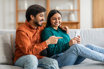 Obraz premium Indian man and woman are seated on a modern couch, engrossed in looking at a cell phone screen. They appear focused and engaged with the device, possibly browsing or communicating.
