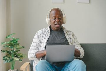 Elderly man with headphones using a laptop on the couch. Comfortable home setting with a plant in the background.