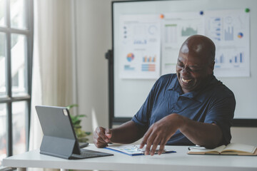 Smiling businessman working on financial report with laptop and documents in office, charts and graphs on whiteboard in background.