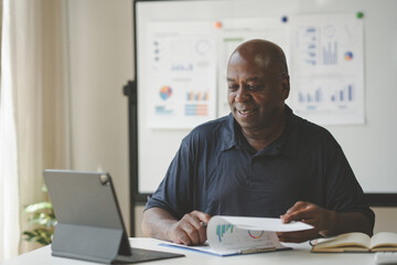 Fototapeta premium Smiling professional working remotely from home, using a tablet for video conferencing, with charts and graphs in the background.