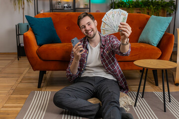 Obraz premium Successful rich man holding smartphone and waving dollar bill money fan sitting on room floor at home. Happy Caucasian guy winning online lottery game planning vacation calculating budget concept
