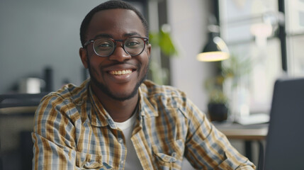 In a bright and modern office space, a young professional with glasses and a flannel shirt smiles warmly while working at his laptop.