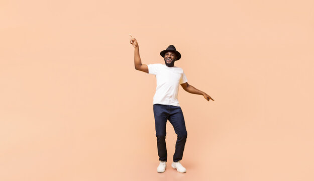 Stylish young African American guy wearing hat dancing, having fun over beige studio background, panorama with copy space