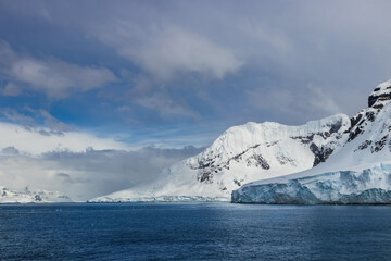 Shoreline, Antarctic Peninsula. Snow, ice, mountains. Blue ocean in the foreground; blue ice visible on shore. Clouds, sky above.
