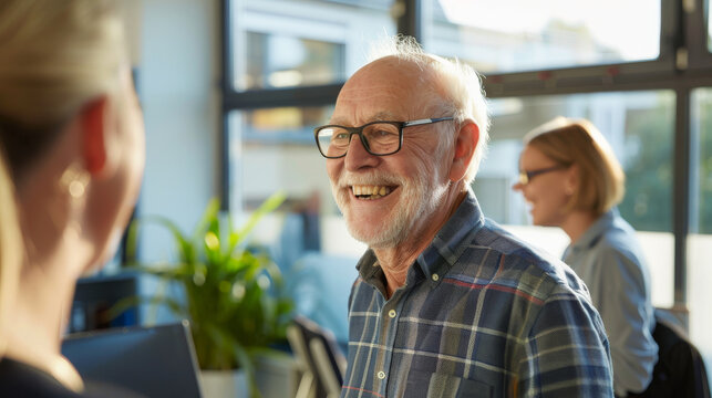 A cheerful elderly man with a white beard and glasses enjoys a lively conversation in a bright, modern office space with colleagues.