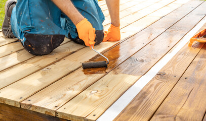 A person kneeling on a wooden deck applies a coat of wood stain with a roller. The wood is mostly...