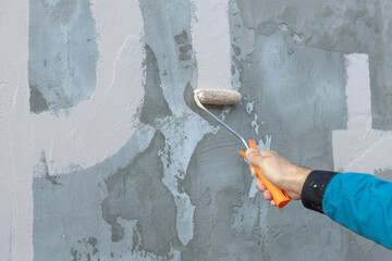 A close-up image of a hand guiding a paint roller across a rough, textured wall, applying a fresh...