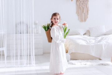 Little girl in a white dress stands in a bright, airy bedroom holding a basket of tulips. The serene setting features white decor, creating a peaceful and gentle atmosphere.