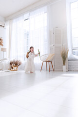 Happy little girl in flowing white dress runs barefoot in bright, sunlit room. Airy space, interior decorated in minimalist style, enhances feeling of freedom, joy. Happiness childhood. Boho style
