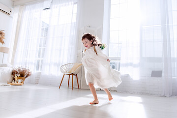 Happy little girl in flowing white dress runs barefoot in bright, sunlit room. Airy space, interior decorated in minimalist style, enhances feeling of freedom, joy. Happiness childhood. Boho style