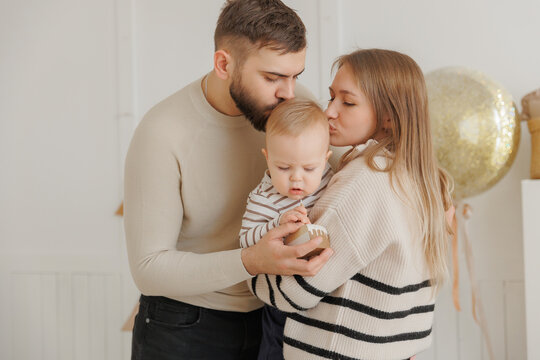 Happy baby boy receives kisses from his loving parents, creating heartwarming scene during first birthday celebration at home. Lifestyle moment of scandinavian family