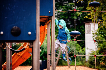 Young child wearing a teal and blue raincoat, climbing on a playground climbing frame on a rainy day. The scene captures the spirit of adventure and play.