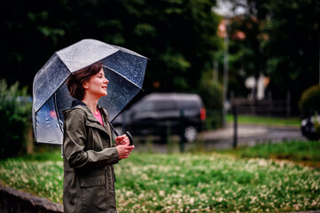 Young woman wearing a pink top and green jacket, smiling while holding a transparent umbrella on a rainy day. The scene captures a moment of joy and serenity.