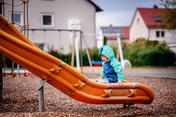 Young child in a teal and blue raincoat, climbing up an orange slide at a playground on a rainy...