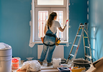 Young woman is doing renovation by herself inside a room with tools and other stuff on the floor. Long shot from behind