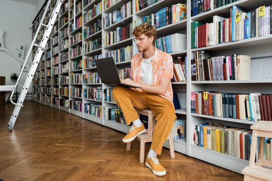 A young man immerses himself in study, typing on a laptop in a library chair.
