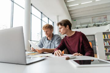 Two young men focus on laptop screen, engrossed in study session at table.