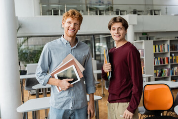 Two young male students stand side by side, each holding a book in a library.