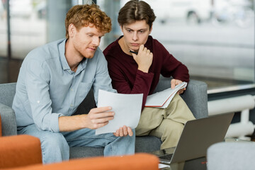 Two men focus on a paper while sitting on a couch.