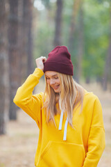 Blue-eyed blonde in a yellow hoodie and burgundy hat in a pine forest. Portrait of a joyful young woman enjoying in autumn park.