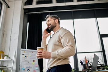 A handsome businessman with a beard is talking on his phone while holding a cup of coffee in a modern office setting.