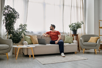 A bearded man in casual attire works remotely from his home office, sitting on a couch with a laptop and a cup of coffee.