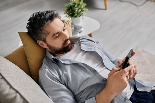 A bearded man in casual attire is lying on a couch, working remotely from home using a smartphone.