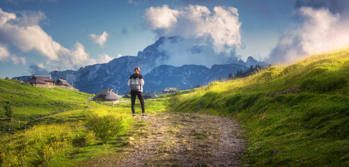 Fototapeta premium Girl on the rural road with yellow flowers and green grass in beautiful alpine mountain valley at sunset in summer. Young woman in old alpine village, sky with clouds. Travel and Hiking. Slovenia