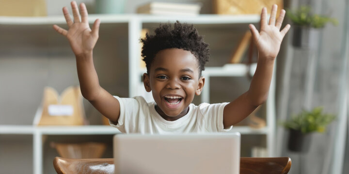 Excited african american school boy raises hands while using laptop