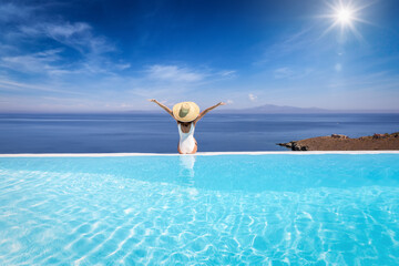 Summer holiday concept with a happy woman with sunhat sitting at the edge of a big infinity pool with view