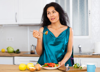 Portrait of asian woman standing at table in kitchen and eating just cooked salad. Woman having breakfast with fresh salad.