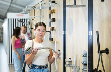 Young female shopper, wearing casual white t-shirt and jeans, carefully choosing new showerhead in...