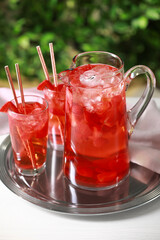 Tasty strawberry lemonade in jug and glasses on white table against blurred green background