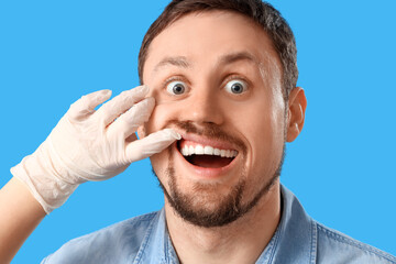 Surprised young man and dentist's hand on blue background, closeup