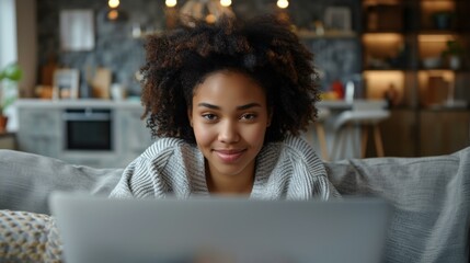 Charming biracial young woman with curly hair smiling while working on laptop in cozy modern apartment with blurred background.