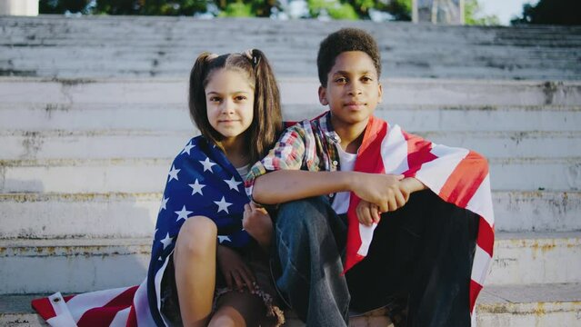 Two young children sit wrapped in an American flag, symbolizing patriotism and unity. They are seated on outdoor steps, looking directly at the camera with a solemn expression.