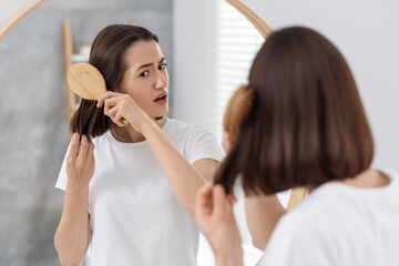 Stressed woman brushing her hair near mirror indoors. Alopecia problem