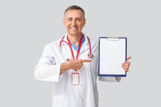 Mature doctor pointing at clipboard on light background