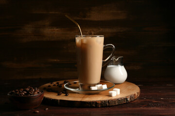 Glass cup of iced latte with coffee beans and jug of milk on wooden background