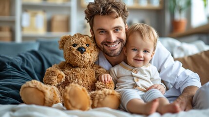 A man and a baby are sitting on a bed with a teddy bear between them. The man is smiling and the baby is smiling as well