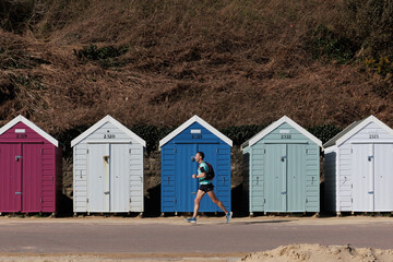 Active sport man running on seafront in beach with row of wooden beach huts on background in spring in Bournemouth, UK. South coast of England.