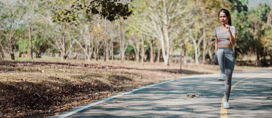 Fototapeta premium Young woman in athletic clothing performing high knees on a paved path in a park surrounded by trees on a sunny day.